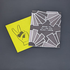 Greeting card and envelope by Amador Collective. Lime Green envelope features an image of a hand giving the peace sign and holding a heart. Card features a black background with white outlined geometric shapes in the corners and reads "before you, my life was black + white" in white hand written letters available at Coyote Supply Co, a zero waste witch store in Midtown Reno, Nevada that is BIPOC owned