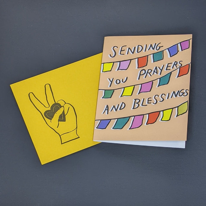 Greeting card and envelope by Amador Collective. Yellow envelope features a hand giving  a peace sign and holding a heart. Card features a tan background with three rows of colorful banners and reads 