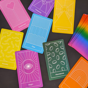 Image features a spread of cards from the Prism Oracle Deck by Nicole Pivirotto. From left to right: Light pink "sweetness," bright purple "creativity," bright yellow "determination,"  rainbow gradient card backs, acid yellow "anxiety," fuchsia "magic," bright blue "flow," dark green "abundance," bright orange card with label not visible in frame.