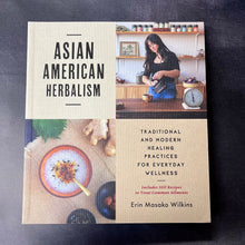 Load image into Gallery viewer, Paperback book cover with beige squares and a photo of a woman pouring tea and a photo of a bowl surrounded by herbs.
The cover reads:
Asian American Herbalism 
Traditional and modern healing practices for everyday wellness
Includes 100 recipes to treat common ailments
Erin Masako Wilkins

This item is sold at Coyote Supply Co a BIPOC and LGBTQIA+ owned zero waste witch store in Reno Nevada