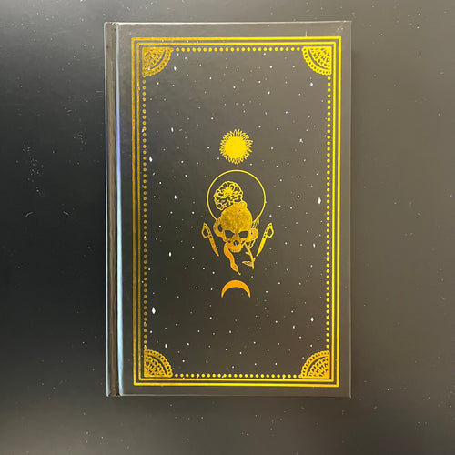 A black hardcover book with gold foil embellishments, the cover has no text only a golden skull surrounded by a marigold, a knife, a rose, a moon, and a sun. Sold at Coyote Supply Co a BIPOC and LGBTQIA+ owned zero waste witch store in Reno Nevada