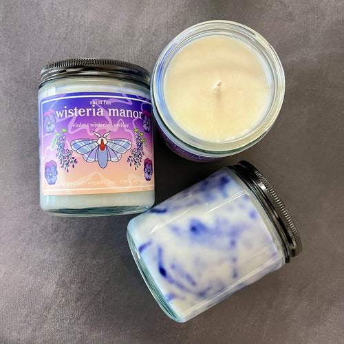 Three candles on a black background. One candle is placed wick up, another is back up showing the purple marbling in the wax, the final candle is label up. The label is purple and champagne with illustrations of a tolype moth surrounded by violets and wisteria vines. The label reads: saint fax
wisteria manor
violet wisteria vetiver
hand poured vegan 8oz 100% soy wax