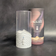 A white colored saalt menstrual disc steamer next to it's packaging cylinder on a dark background. The dark colored packaging cylinder has white text that reads:
Sanitize cups and discs of all sizes
Saalt
menstrual cup & disc steamer
Period care simplified
BPA-Free

This item is sold at Coyote Supply Co a BIPOC and LGBTQIA+ owned zero waste witch store in Reno Nevada