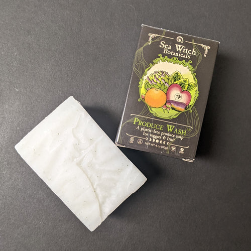 A white bar of canary clean next to it's black box on a black background. The box has an illustration of various fruits and veg in a green circular frame (oranges, carrots, apples, radishes) the label reads: Sea Witch Botanicals 
PRODUCE WASH
A plastic-free produce soap for veggies & fruit