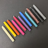 A rainbow of chime candles arranged diagonally across the frame from white, yellow, orange, pink, red, purple, blue, green, brown, black, to grey on a black background. 
Sold at Coyote Supply Co a BIPOC and LGBTQIA+ owned zero waste witch store in Reno Nevada