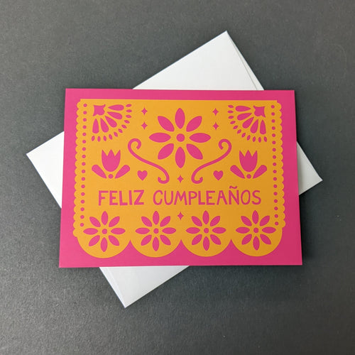 A pink card with an orange papel picada design that reads Feliz Cumpleanos. Sold at Coyote Supply Co a BIPOC and LGBTQIA+ owned zero waste witch store in Reno Nevada 