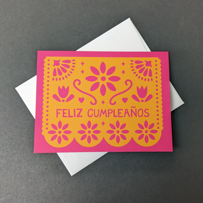 A pink card with an orange papel picada design that reads Feliz Cumpleanos. Sold at Coyote Supply Co a BIPOC and LGBTQIA+ owned zero waste witch store in Reno Nevada 