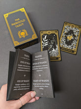 Load image into Gallery viewer, Black background with tarot deck scattered on it and a hand in the lower left corner. Cards, box, and guide book are gold and white skeletons and botanicals on black paper. Sold at Coyote Supply Co a BIPOC and LGBTQIA+ owned zero waste witch store in Reno Nevada