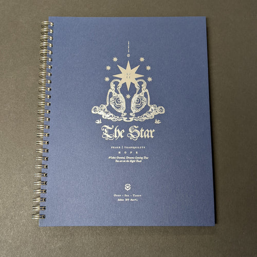 A blue spiralbound journal with silver foil details on the cover. The cover reads The Star 
peace tranquility 
hope
There is a illustration of a lage star surrounded by smaller stars and two ornate vases. 
Sold at Coyote Supply Co a BIPOC and LGBTQIA+ owned zero waste witch store in Reno Nevada