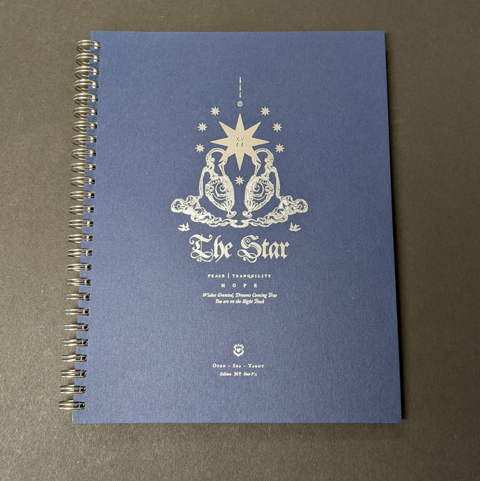 A blue spiralbound journal with silver foil details on the cover. The cover reads The Star 
peace tranquility 
hope
There is a illustration of a lage star surrounded by smaller stars and two ornate vases. 
Sold at Coyote Supply Co a BIPOC and LGBTQIA+ owned zero waste witch store in Reno Nevada