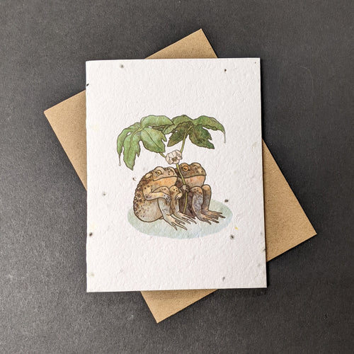 A white seed paper card with a brown craft envelope behind it on a black background. The card has a watercolor style illustration of two toads sitting together under large leaves.

Sold at Coyote Supply Co a BIPOC and LGBTQIA+ owned zero waste witch store in Reno Nevada