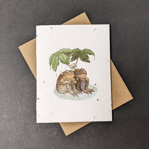 A white seed paper card with a brown craft envelope behind it on a black background. The card has a watercolor style illustration of two toads sitting together under large leaves.

Sold at Coyote Supply Co a BIPOC and LGBTQIA+ owned zero waste witch store in Reno Nevada