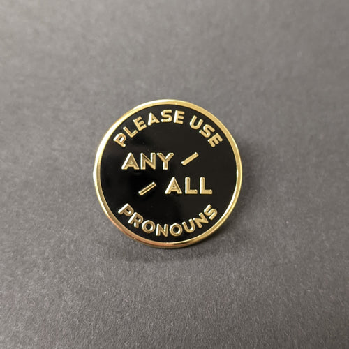 A circular black enamel pin with gold text on a black background. The pin reads:
PLEASE USE ANY/ALL PRONOUNS

Sold at Coyote Supply Co a BIPOC and LGBTQIA+ owned zero waste witch store in Reno Nevada