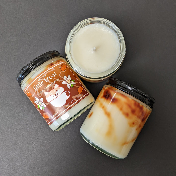 Three candles on a black background. One candle is placed wick up, another is back up showing the brown marbling in the wax, the final candle is label up. The label is brown with illustrations of a coffee mug with a foam top that resembles a shiba inu dog surrounded by caramels and vanilla flowers. The label reads: saint fax
little treat
coffee vanilla caramel
hand poured vegan 8oz 100% soy wax