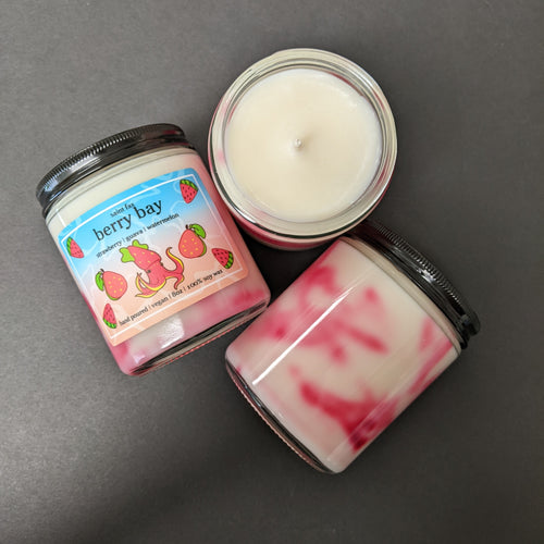 Three candles on a black background. One candle is placed wick up, another is back up showing the red marbling in the wax, the final candle is label up. The label is blue-pink with illustrations of a strawberry squid surrounded by pink guava and watermelon slices. The label reads: saint fax
berry bay
strawberry guava watermelon
hand poured vegan 8oz 100% soy wax