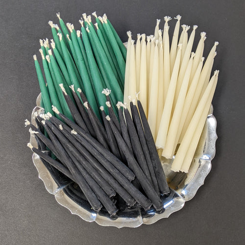 A round silver plate overflowing with small candles arranged in three groups laid on the plate. The groups are black candles, white candles, then green candles. 
Sold at Coyote Supply Co a BIPOC and LGBTQIA+ owned zero waste witch store in Reno Nevada