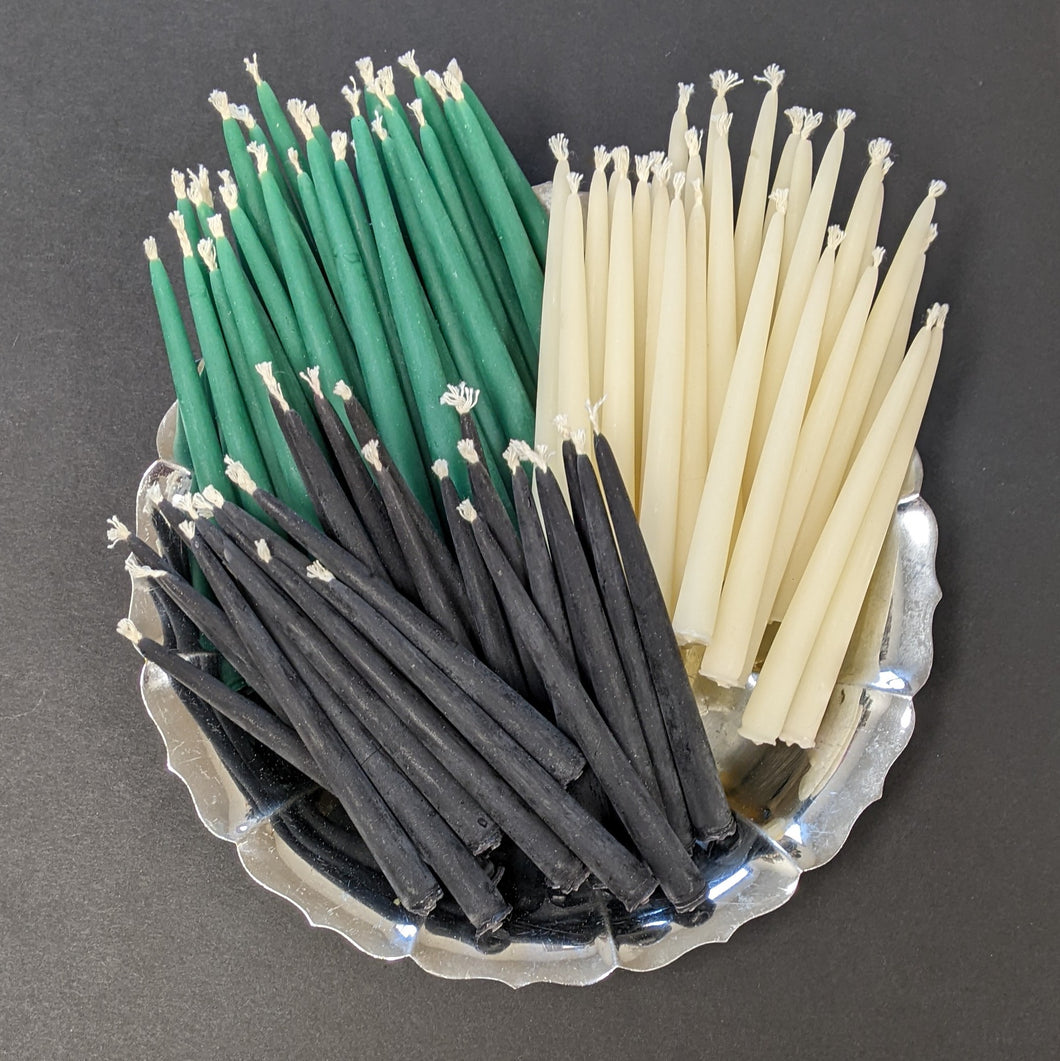 A round silver plate overflowing with small candles arranged in three groups laid on the plate. The groups are black candles, white candles, then green candles. 
Sold at Coyote Supply Co a BIPOC and LGBTQIA+ owned zero waste witch store in Reno Nevada