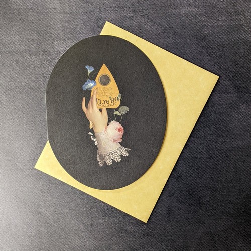 A black rounded greeting card with and illustration of  a hand holding a planchette surrounded by flowers. With a yellow envelope. 
Sold at Coyote Supply Co a BIPOC and LGBTQIA+ owned zero waste witch store in Reno Nevada