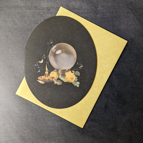 A rounded black greeting card with an illustration of a crystal ball, a candle, a small frog, and yellow roses. Behind the card is a yellow envelope. 
Sold at Coyote Supply Co a BIPOC and LGBTQIA+ owned zero waste witch store in Reno Nevada
