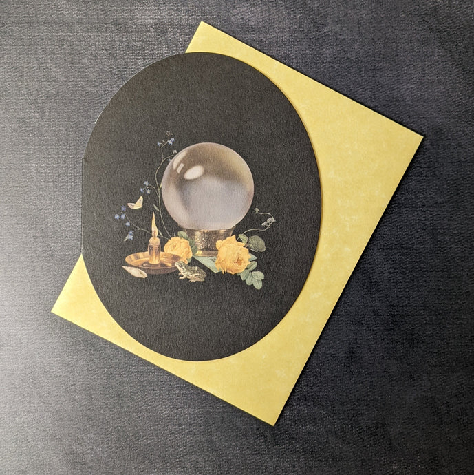 A rounded black greeting card with an illustration of a crystal ball, a candle, a small frog, and yellow roses. Behind the card is a yellow envelope. 
Sold at Coyote Supply Co a BIPOC and LGBTQIA+ owned zero waste witch store in Reno Nevada