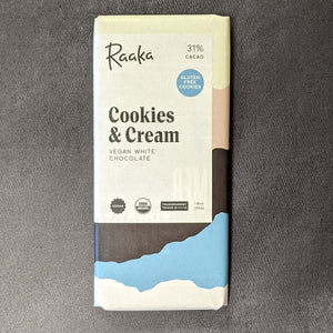 A paper wrapped chocolate bar, the paper has a white square with black text and colors from top to bottom of white, grey, black, blue, and white. The label has black text that reads "Raaka 31% CACAO
GLUTEN FREE COOKIES
Cookies & Cream 
VEGAN WHITE CHOCOLATE"
Sold at Coyote Supply Co a BIPOC and LGBTQIA+ owned zero waste witch store in Reno Nevada