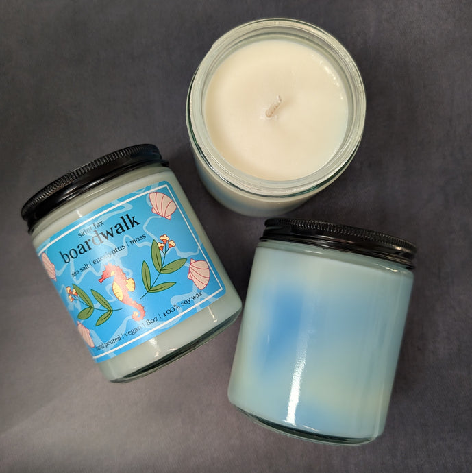 Three candles on a black background. One candle is placed wick up, another is back up showing the blue marbling in the wax, the final candle is label up. The label is light blue with illustrations of a pacific sea horse surrounded by eucalyptus leaves and sea shells. The label reads: saint fax
boardwalk
sea salt eucalyptus moss
hand poured vegan 8oz 100% soy wax