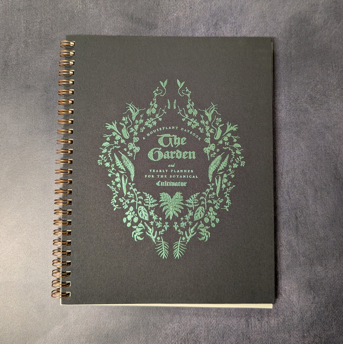 A spiral bound black journal with green foil details. The cover reads: A houseplant  daybook 
The Garden
and
yearly planner for the botanical cultivator.
The text is surrounded by illustrations of plants and leaves
Sold at Coyote Supply Co a BIPOC and LGBTQIA+ owned zero waste witch store in Reno Nevada