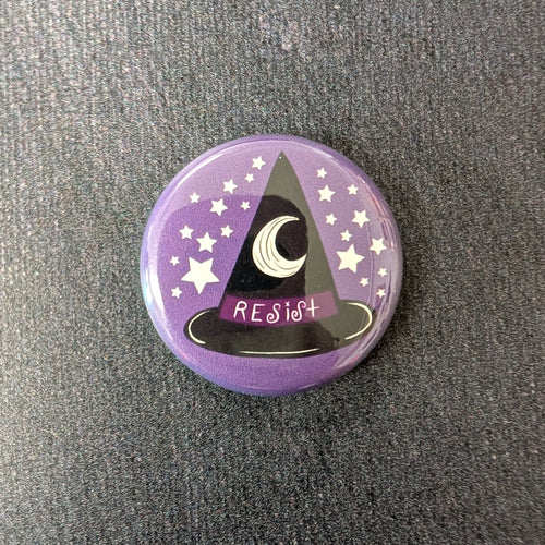 A glossy purple button with white stars and a black witch hat with a dark purple band illustration. The band of the at says 