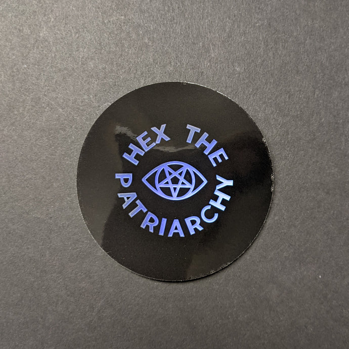 A circular black glossy sticker with holographic text in a circle around a holographic eye with a pentagram iris that reads: Hex the Patriarchy. Sold at Coyote Supply Co a BIPOC and LGBTQIA+ owned zero waste witch store in Reno Nevada