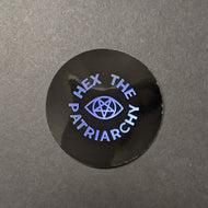 A circular black glossy sticker with holographic text in a circle around a holographic eye with a pentagram iris that reads: Hex the Patriarchy. Sold at Coyote Supply Co a BIPOC and LGBTQIA+ owned zero waste witch store in Reno Nevada