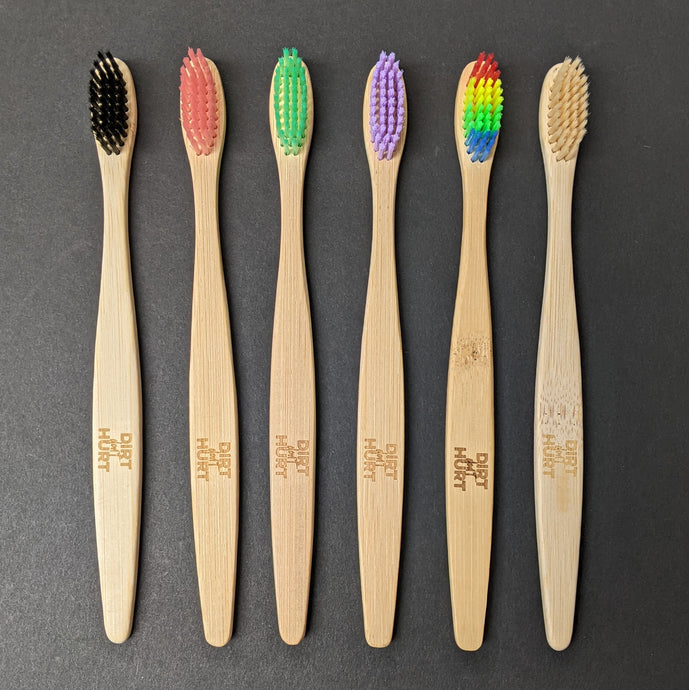 Six bamboo tooth brushes with Dirt Don't Hurt brandng on the handle. The colors of each brush from left to right are black, pink, green, purple, rainbow, and beige. Sold at Coyote Supply Co a BIPOC and LGBTQIA+ owned zero waste witch store in Reno Nevada