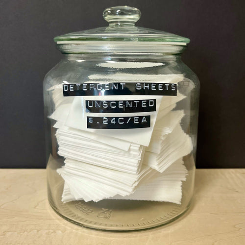 A glass apothocary style jar with a stack of white rectangular detergent sheets. The jar has black labels on the front that read: Detergent Sheets Unscented $ .24c / EA. Sold at Coyote Supply Co a BIPOC and LGBTQIA+ owned zero waste witch store in Reno Nevada 