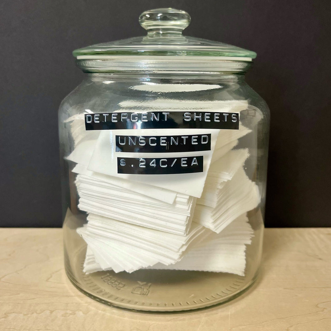 A glass apothocary style jar with a stack of white rectangular detergent sheets. The jar has black labels on the front that read: Detergent Sheets Unscented $ .24c / EA. Sold at Coyote Supply Co a BIPOC and LGBTQIA+ owned zero waste witch store in Reno Nevada 