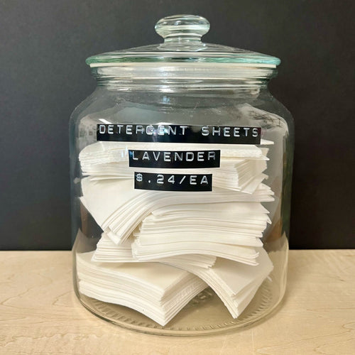 A glass apothocary style jar with a stack of white rectangular detergent sheets. The jar has black labels on the front that read: Detergent Sheets Lavender $ .24c / EA. Sold at Coyote Supply Co a BIPOC and LGBTQIA+ owned zero waste witch store in Reno Nevada 