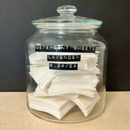 A glass apothocary style jar with a stack of white rectangular detergent sheets. The jar has black labels on the front that read: Detergent Sheets Lavender $ .24c / EA. Sold at Coyote Supply Co a BIPOC and LGBTQIA+ owned zero waste witch store in Reno Nevada 