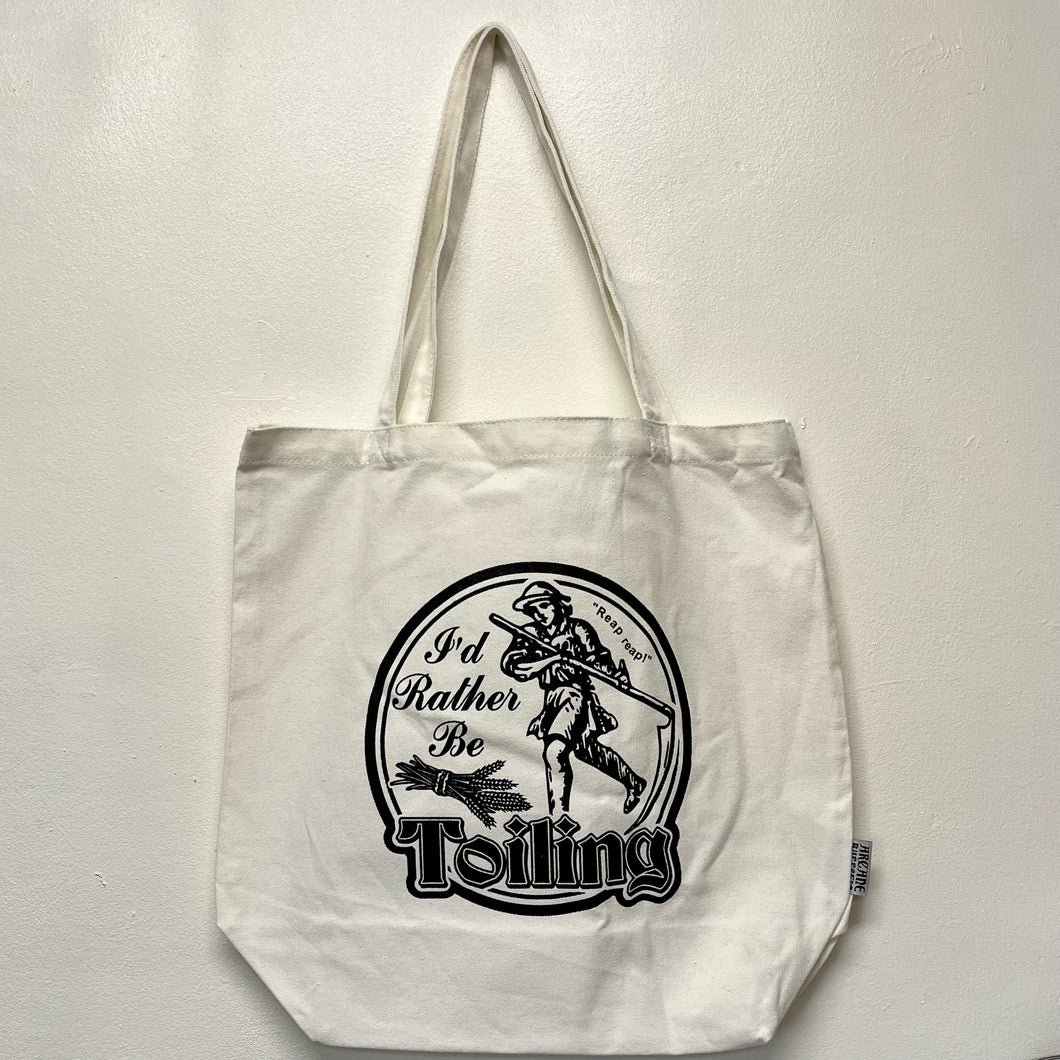 A white tote bag with a black screen printed design of a medieval peasant that reads I'd Rather Be Toiling.  Sold at Coyote Supply Co a BIPOC and LGBTQIA+ owned zero waste witch store in Reno Nevada 