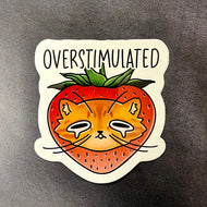 A sticker of a cartoon orange cat head in a strawberry with black text pictured on a dark background.
The text reads:
OVERSTIMULATED

Sold at Coyote Supply Co a BIPOC and LGBTQIA+ owned zero waste witch store in Reno Nevada
