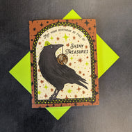 A brown greeting card with a checkered frame and a raven with a disco ball in its mouth. Around the raven is black text that reads: 
May your birthday be full of 
Shiny
Treasures

Sold at Coyote Supply Co a BIPOC and LGBTQIA+ owned zero waste witch store in Reno Nevada 
