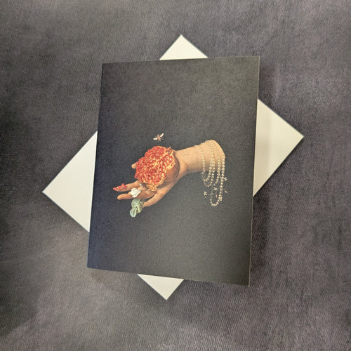 A black card with an illustration of a hand with pearl bracelets holding a half open pomegranate. 
Also includes white envelope.
Sold at Coyote Supply Co a BIPOC and LGBTQIA+ owned zero waste witch store in Reno Nevada