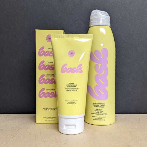 From left to right  a yellow box that reads Bask, sheer, vegan, uva/uvb protection sunscreen broad spectrum SPF 50 lotion. A yellow tube of sunscreen that reads Bask, sheer, moisturizing sunscreen, broad spectrum SPF 50 lotion, water & sweat resistant. A yellow bottle of sunscreen that reads Bask, non-aerosol invisible mist sunscreen, broad spectrum SPF 50 continuous spray, water & sweat resistant. Sold at Coyote Supply Co a BIPOC and LGBTQIA+ owned zero waste witch store in Reno Nevada 