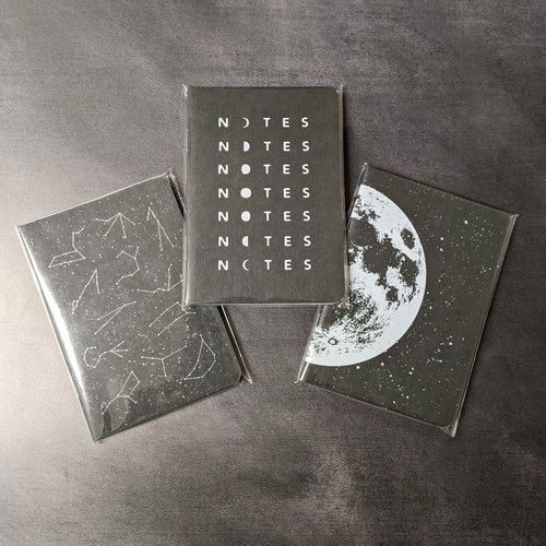 Three black paper cover notebooks on a black background with varying covers. The left notebook has various constellations, the center notebook reads 