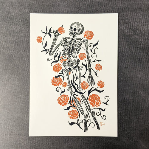 A print of an illustration of a monochrome skeleton surrounded by orange marigolds by Bat In Your Belfry. Sold at Coyote Supply Co a BIPOC and LGBTQIA+ owned zero waste witch store in Reno Nevada 