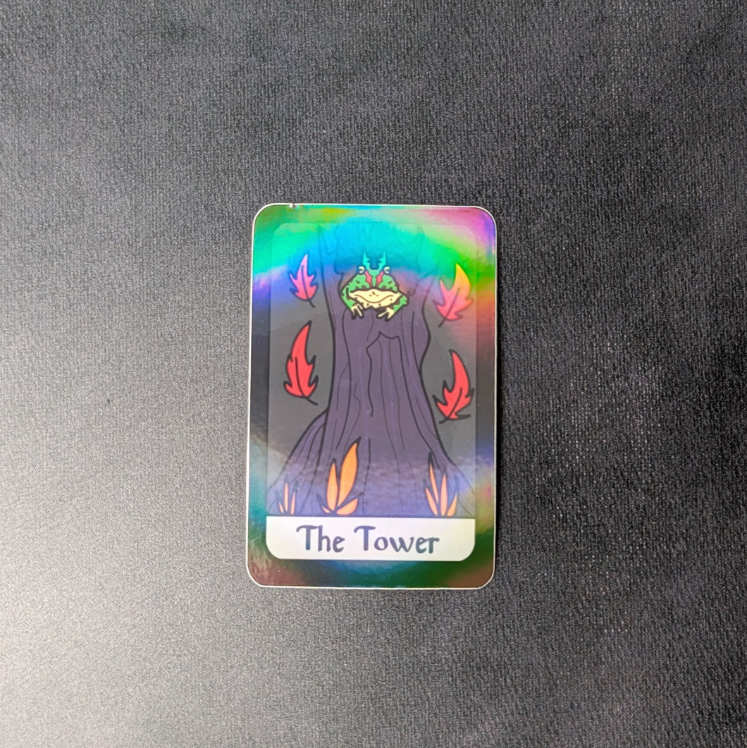 A holographic vinyl sticker of The Tower tarot card. The card illustration is of an Argentinian horned frog in a tower looking tree stump with red leaves falling around him and orange vegetation in the foreground. Sold at Coyote Supply Co a BIPOC and LGBTQIA+ owned zero waste witch store in Reno Nevada 