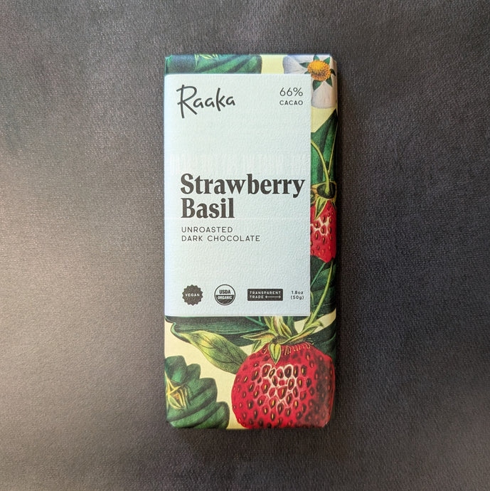 A chocolate bar wrapped in a light yellow paper wrapper with illustrations of strawberry and basil leaves, in the center is a white square with black text. The text reads 