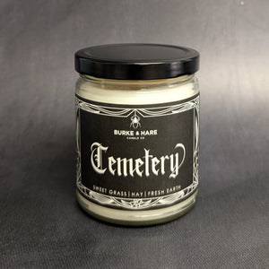 An 8oz vegan candle with a black metal lid and a grey label that reads Burke & Hare Co Cemetary Gates a fresh blend of sweet grass, hay, and earth. Sold at Coyote Supply Co a BIPOC and LGBTQIA+ owned zero waste witch store in Reno Nevada 