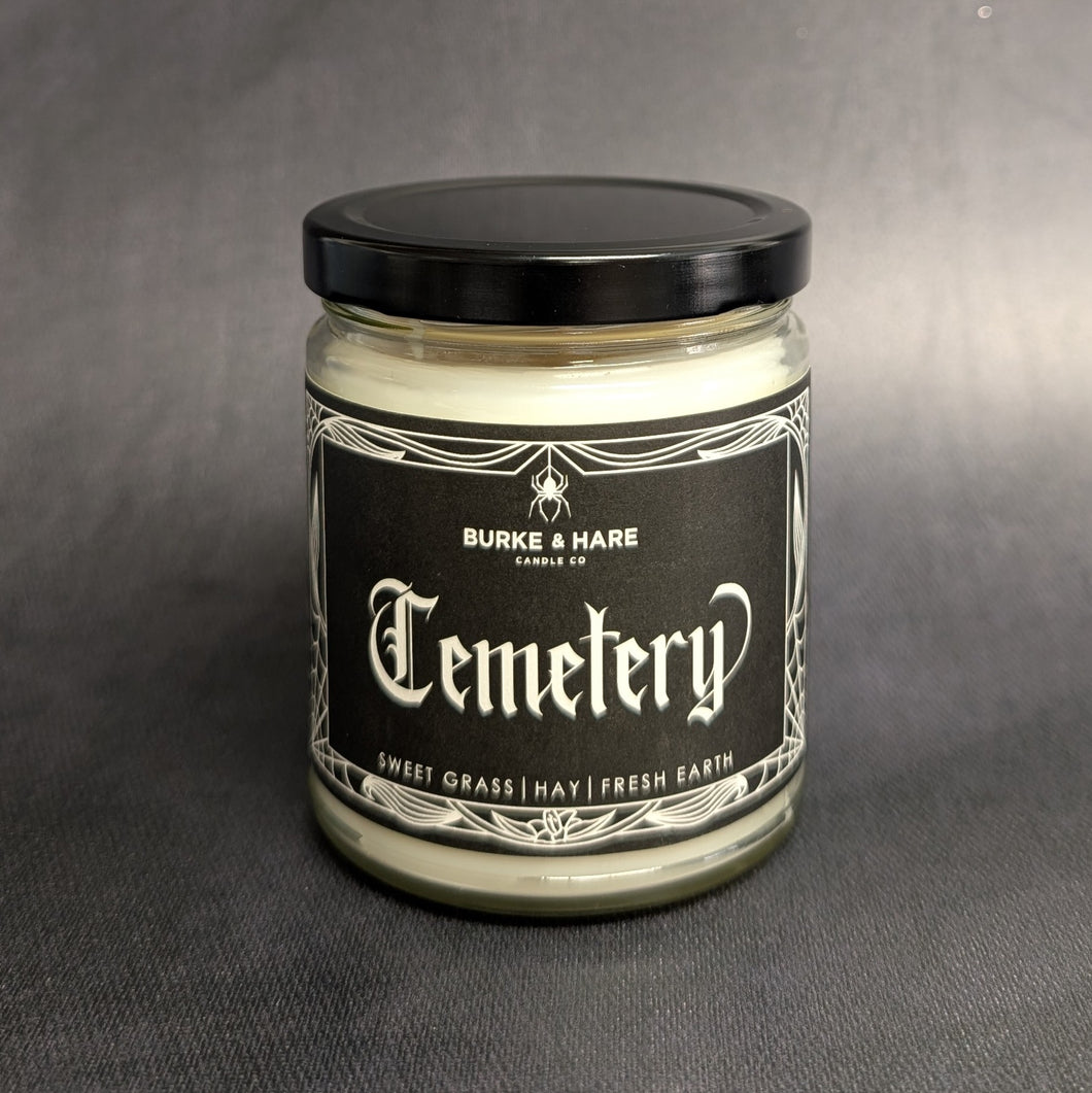 An 8oz vegan candle with a black metal lid and a grey label that reads Burke & Hare Co Cemetary Gates a fresh blend of sweet grass, hay, and earth. Sold at Coyote Supply Co a BIPOC and LGBTQIA+ owned zero waste witch store in Reno Nevada 