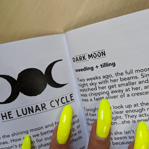 Black text on white paper reads "The Lunar Cycle" & "Dark Moon".  Boss Witch's hand is on the page too & their nails are fluorescent green. 