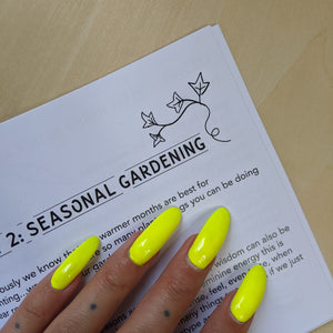 Black text on white paper reads "Seasonal Gardening" with a flourish of ivy in the top right corner.  Boss Witch's hand is on the page too & their nails are fluorescent green. 