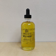Load image into Gallery viewer, Glass bottle with black dropper lid containing golden multi use oil by Motherland Essentials reads &quot;wild rose&quot; in black block letters available at Coyote Supply Co, a zero waste witch store in Midtown Reno, Nevada that is BIPOC owned