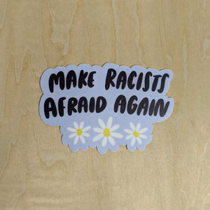 Baby blue sticker with black text reads "make racists afraid again" with white and yellow daisies available at Coyote Supply Co, a zero waste witch store in Midtown Reno, Nevada that is BIPOC owned